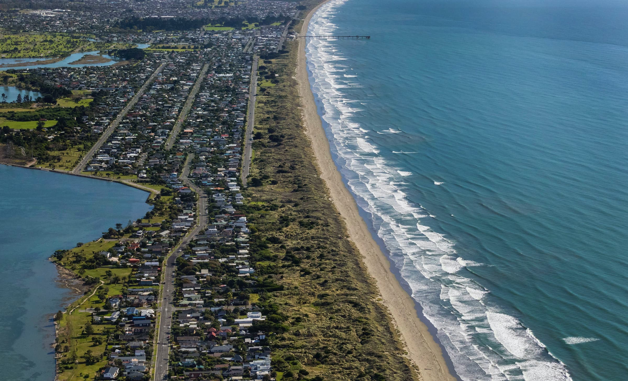 Aerial view of New Brighton Pier and the surrounding coastline, showing the beach, shoreline, parts of New Brighton, and the wider Christchurch area stretching inland