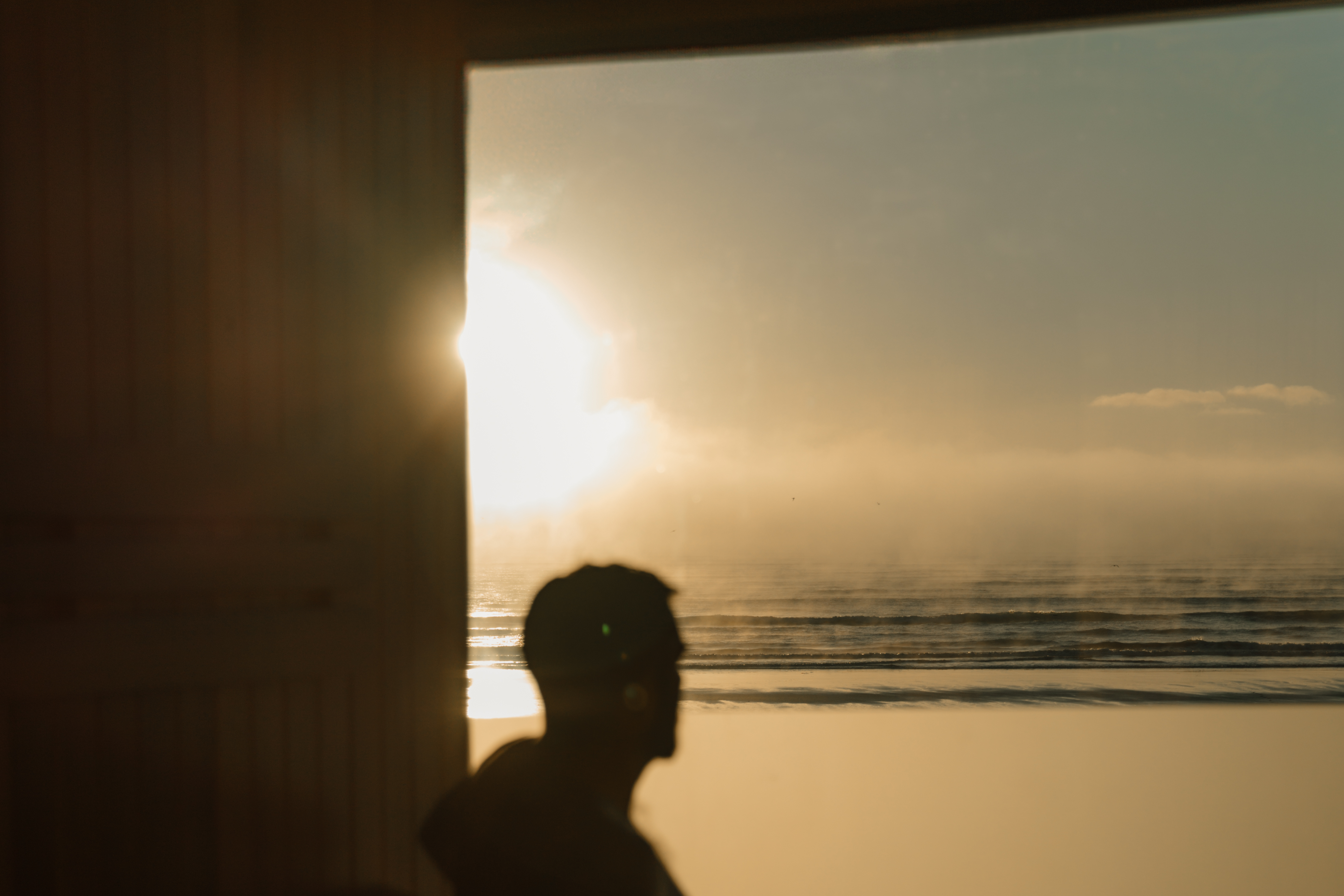 Blurred figure sitting in a sauna, looking out through a window towards the ocean, with the coastal view as the main focus of the image