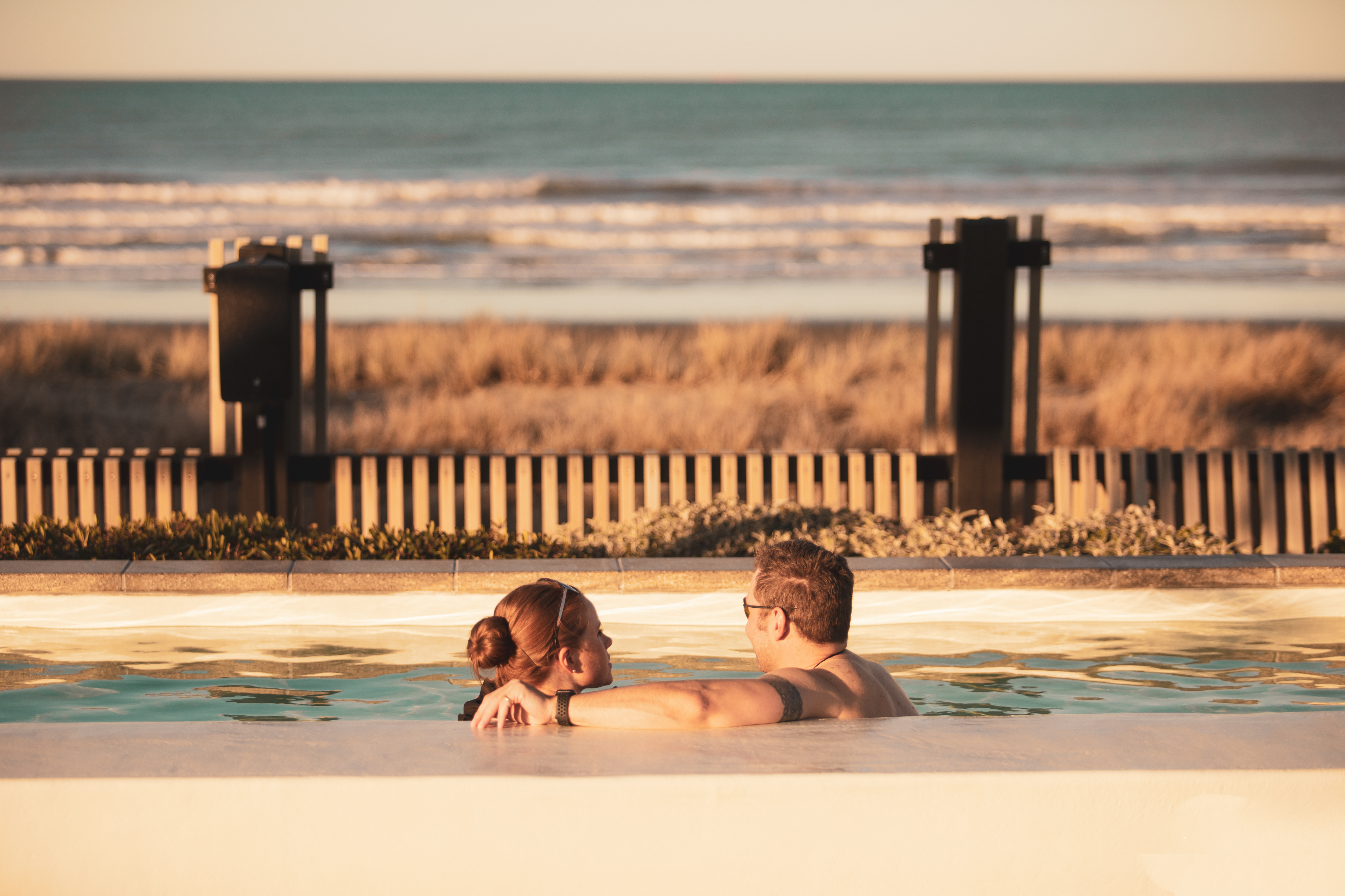A couple relax in a hot pool, looking out over the beach and ocean beyond