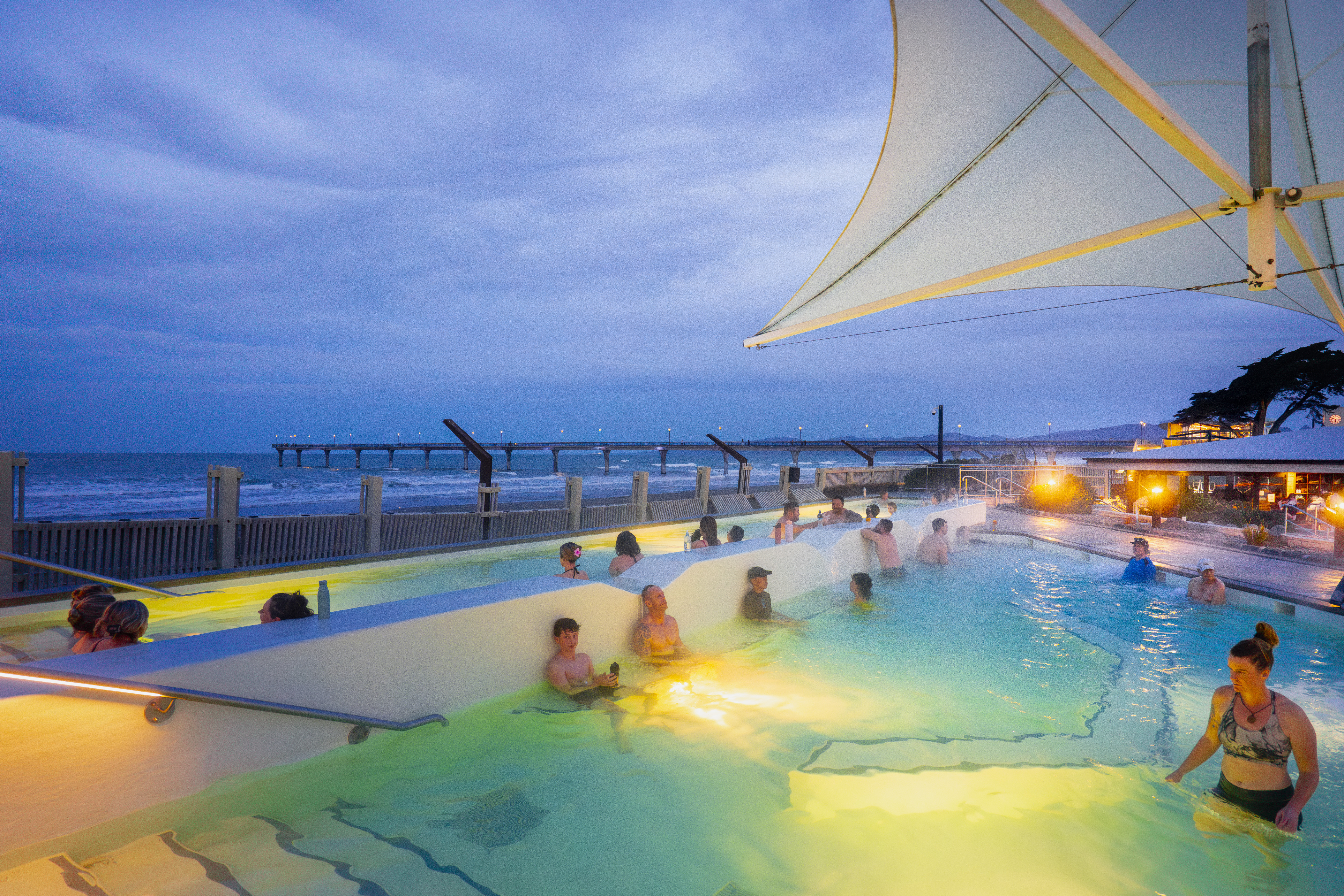 People relaxing in the hot pools during the evening, looking out towards New Brighton Pier and enjoying the coastal views as the sun sets