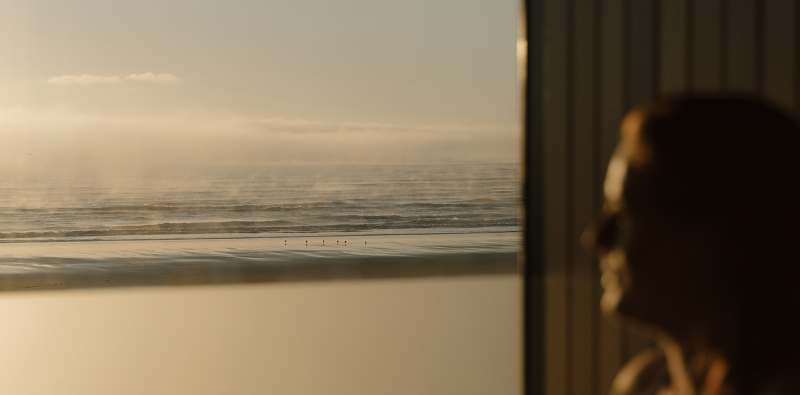 Person sitting in a sauna, softly out of focus in the foreground, with the main view looking through the window to the ocean and coastal scenery beyond