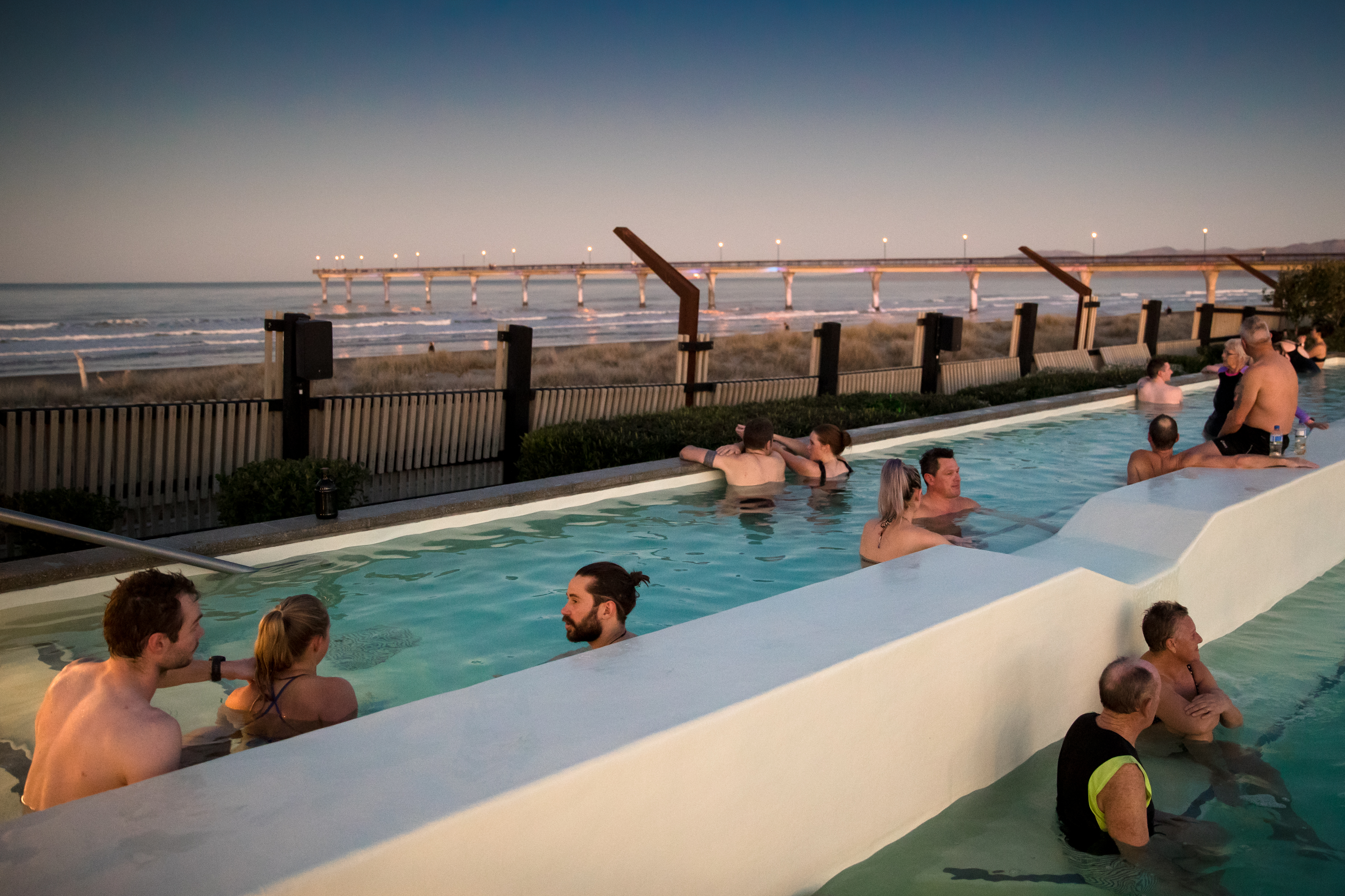 People sit in a hot pool, looking out toward the iconic New Brighton Pier stretching across the ocean