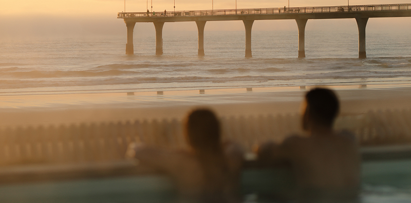 Man and woman relaxing together in hot pools, looking out over New Brighton Pier and the ocean beyond