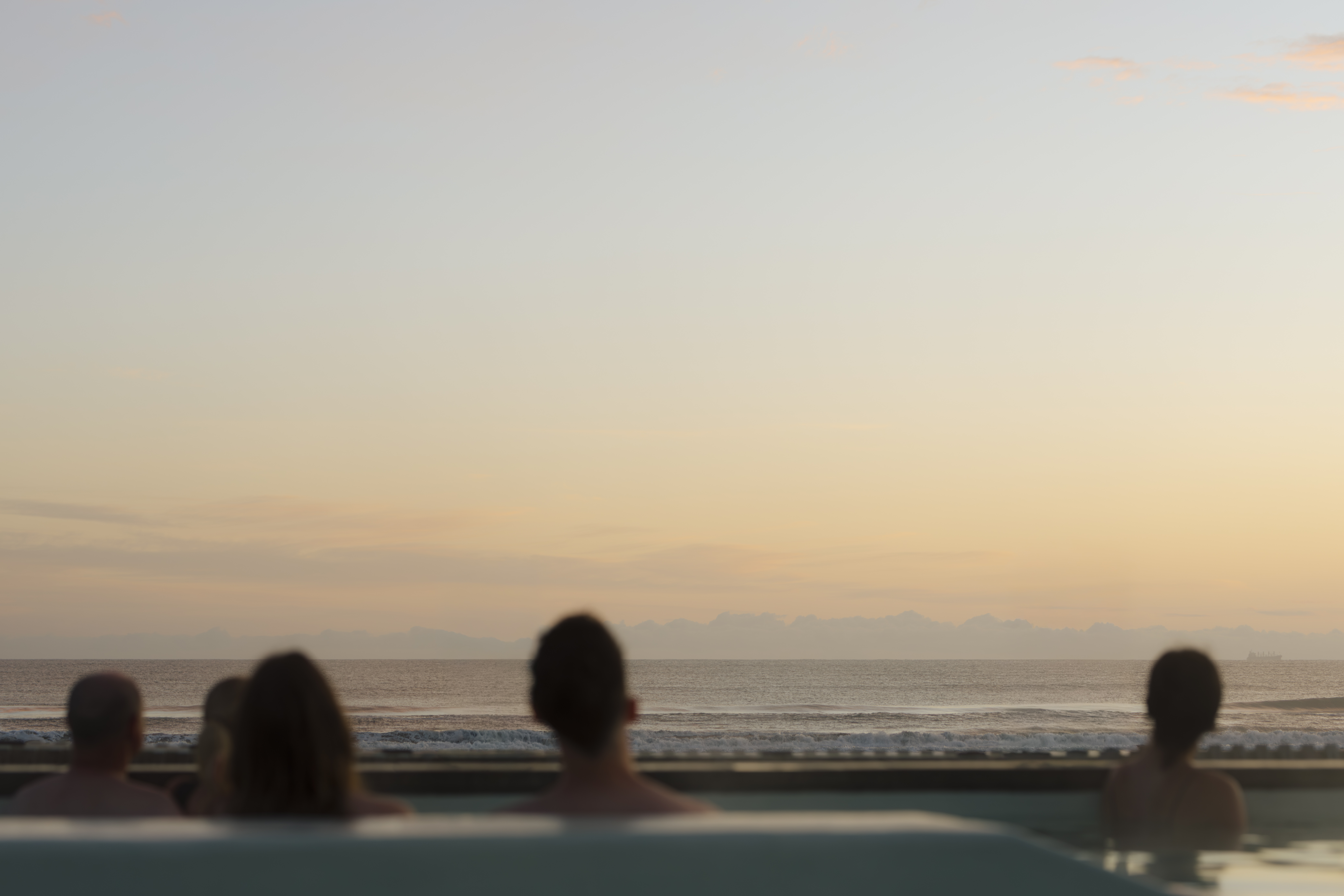Softly blurred heads in the foreground looking out over the ocean, with distant mountains visible on the horizon beneath the open sky