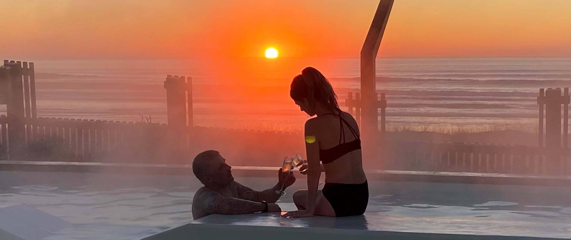 A couple relax in a hot pool at sunrise, smiling as they clink mocktails together