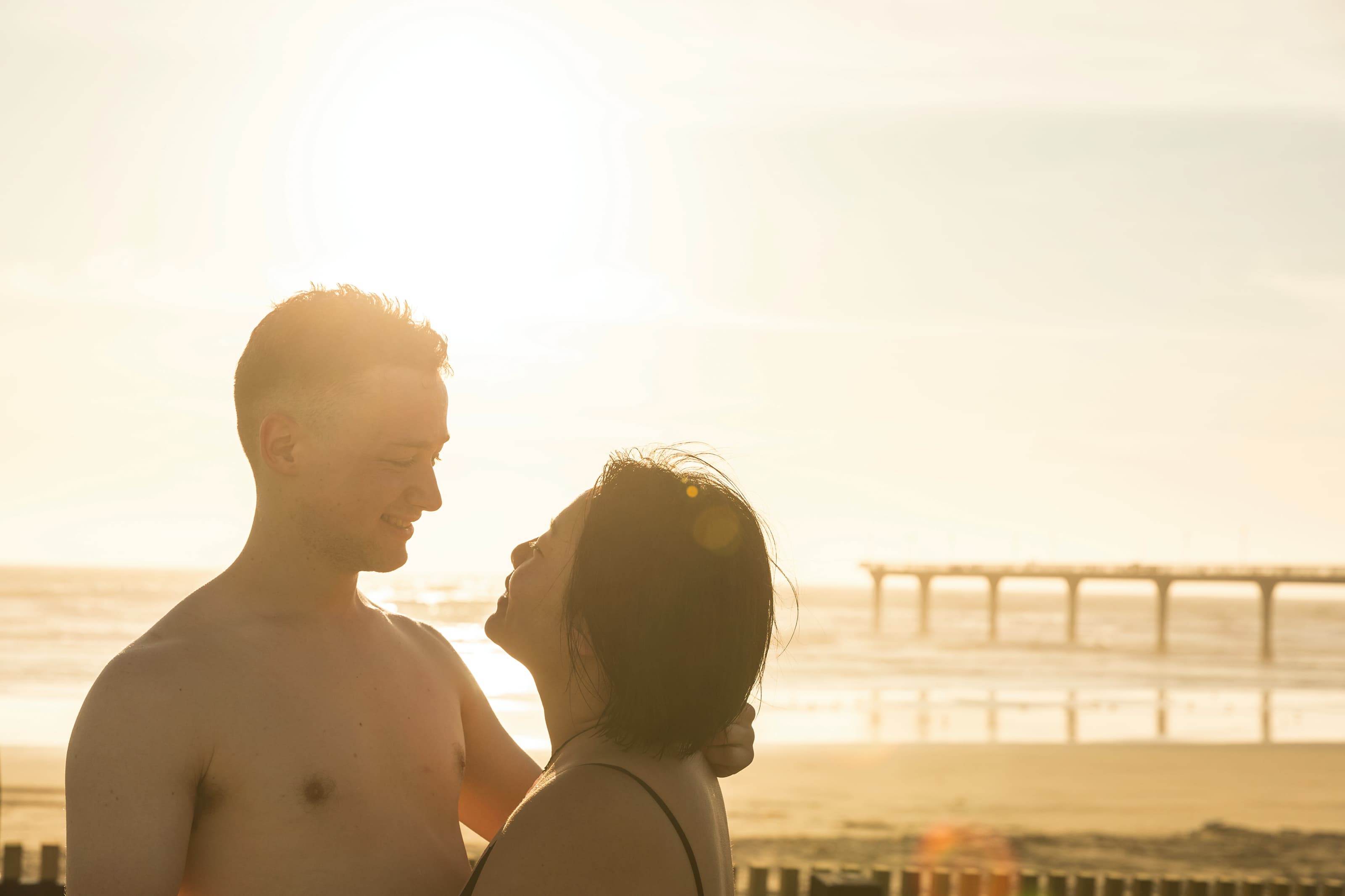 Man and woman relaxing in the hot pools, looking at each other with the beach and New Brighton Pier visible in the background